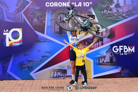 Una ciclista levanta su bicicleta sobre la cabeza junto a un niño de camiseta amarilla, frente al muro «Coroné La 10»
