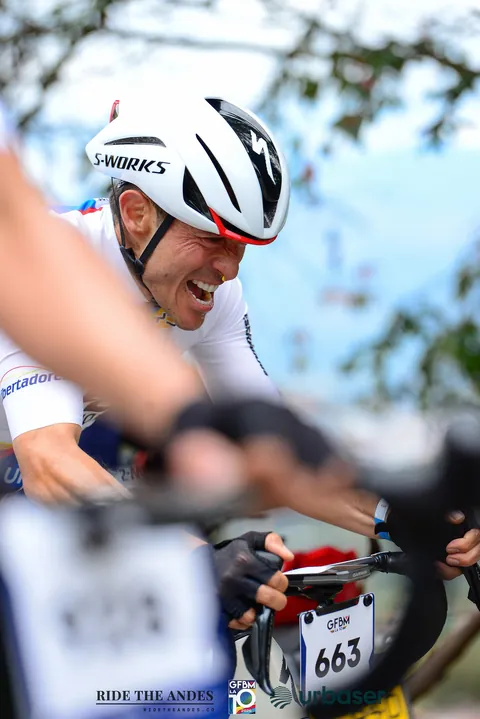 Primer plano de un ciclista que sonríe con todos los dientes; delante, desenfocada, la mano de alguien que lo saluda