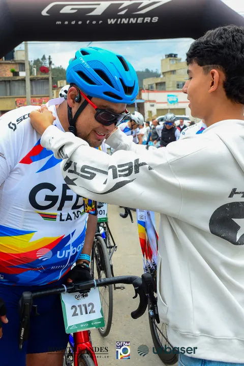 Un joven le pone las manos en los hombros a un ciclista de casco azul, que sonríe, bajo un arco inflable