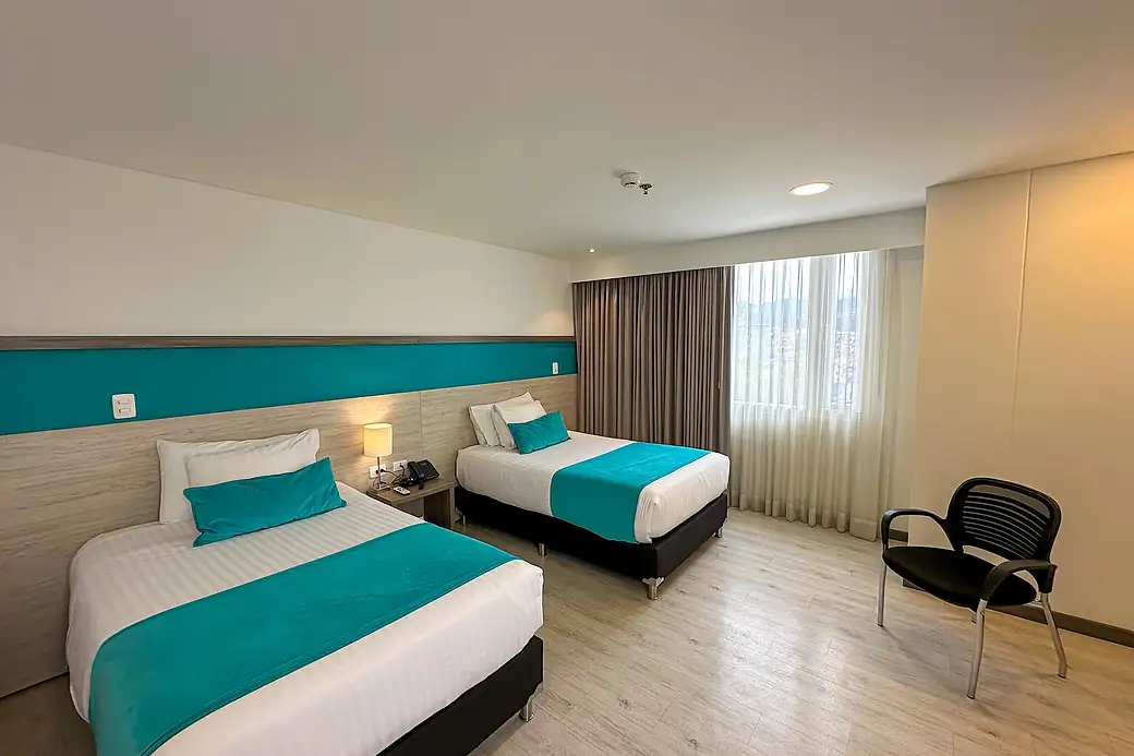 A room with two double beds, a wooden headboard with a turquoise band, a bedside table with a phone and a curtained window.