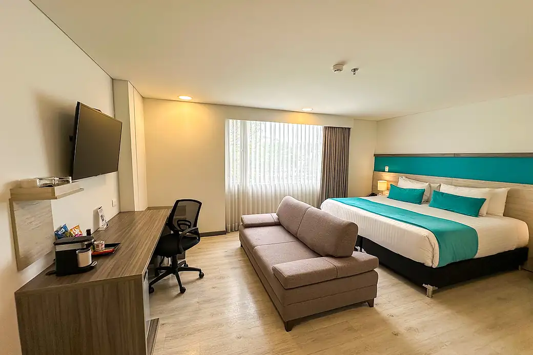 A room with a king bed, a sofa, a desk with an office chair and a wall-mounted television.