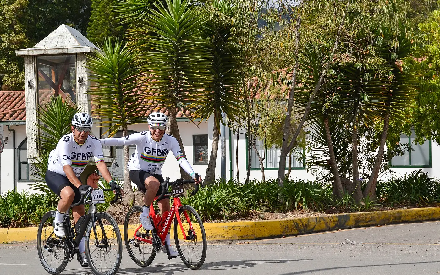 Dos ciclistas ruedan juntos y sonrientes, uno con la mano apoyada en la espalda del otro.