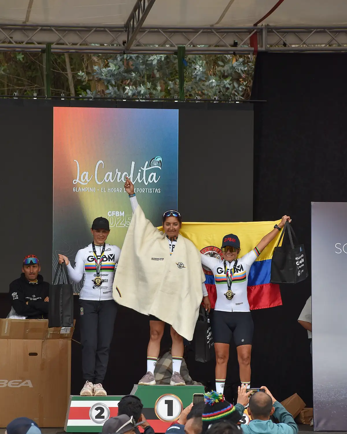 Podio femenino: la ganadora levanta los brazos con una ruana blanca sobre los hombros y la bandera de Colombia detrás.