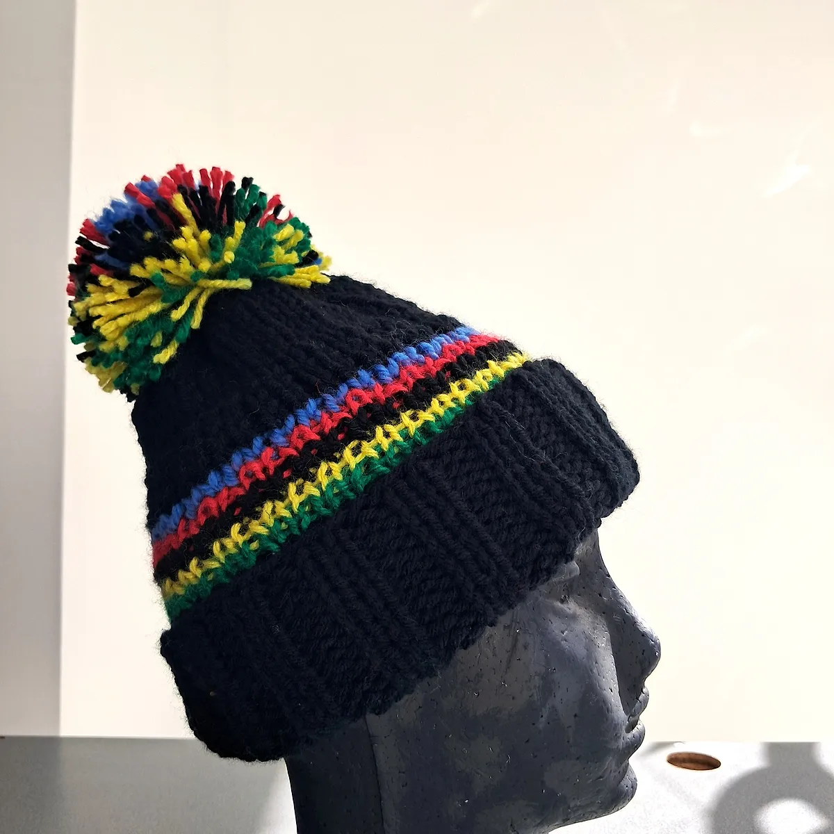 Black wool beanie in profile, with the rainbow bands and the pompom